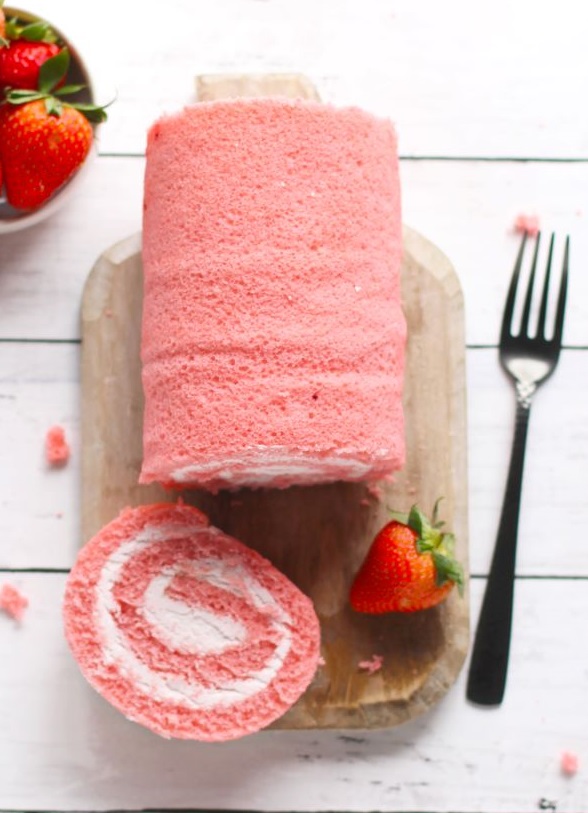 Strawberries & Cream Ambient Cake Roll from River Bend on cutting board, one slice cut off, with a fork, and a bowl of strawberries for garnish.